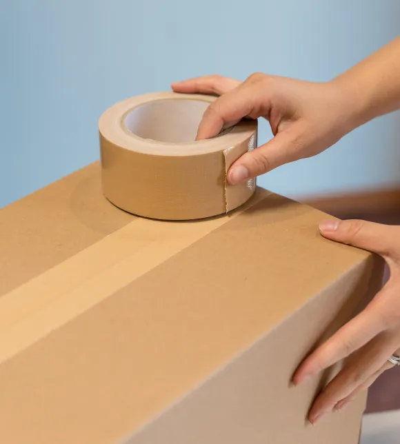引越しサポートでの荷造り作業。茶色の段ボール箱をクラフトガムテープで梱包している人の手元の様子。迅速な作業が伝わる梱包の瞬間。